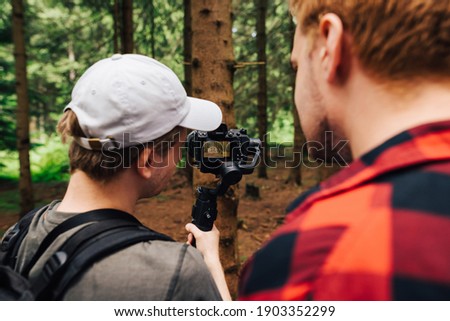 Two men stand on the nature in the woods and watch videos on the erkan filmed on camera with a stabilizer. Backstage photo two operators shoot. content in a coniferous forest. Creating video content.