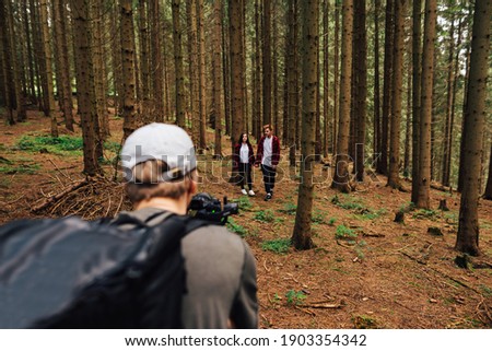 Video cameraman with a camera on a stabilizer in his hands shoots a beautiful young couple man and woman in red shirts walking through the woods holding hands.