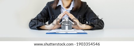 A female home salesperson puts both hands in a triangle pose on a gray miniature house model, Real estate trading ideas. Banner background with copy space.