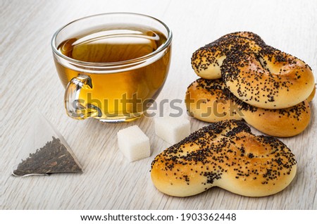 Transparent cup with tea, cookies with poppy seeds, tea bag, sugar cubes on wooden table