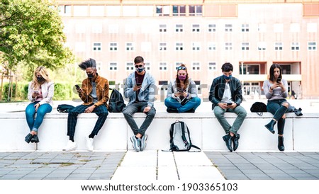 Friends using smart phone covered by mask on Covid third wave - Worried guys and girls watching video news on mobile smartphone - College students sitting at university campus - Focus in central faces Royalty-Free Stock Photo #1903365103