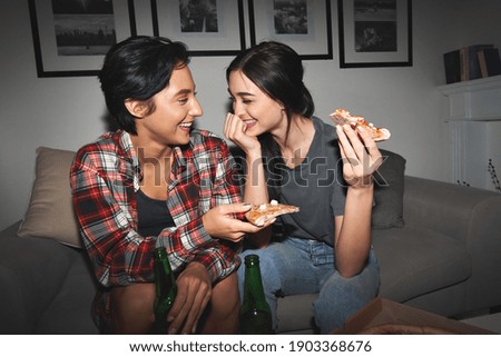 Two happy young girls laughing, drinking beer, sharing pizza at home party late. Cheerful best friends or couple talking, having fun enjoying eating fast food at night sitting on couch together. Royalty-Free Stock Photo #1903368676