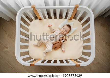 Top view of adorable infant in stylish pajama lying on pillow crown in comfortable cot at home. Baby with teddy bear in cradle in baby's room Royalty-Free Stock Photo #1903375039