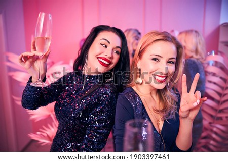 Smiling dark-haired woman looking at a cheerful blonde lady making a V-sign at the party