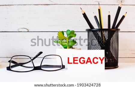 The word LEGACY written on a white business card next to pencils in a stand and glasses. Nearby is a potted plant.