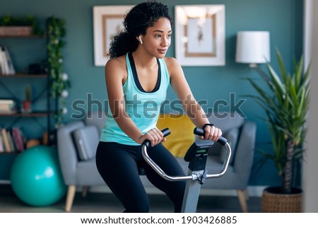 Shot of sporty african young woman exercising on smart stationary bike and listening to music at home. Royalty-Free Stock Photo #1903426885