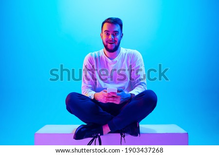 Using cellphone. Caucasian man's portrait with device on blue studio background in neon light. Beautiful male model. Concept of human emotions, facial expression, sales, ad. Copyspace for ad. Close up