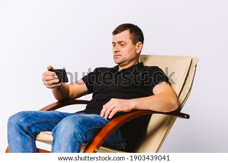 Deaf senior man with a hearing aid sitting in a leather armchair watching a movie on the phone. White background.