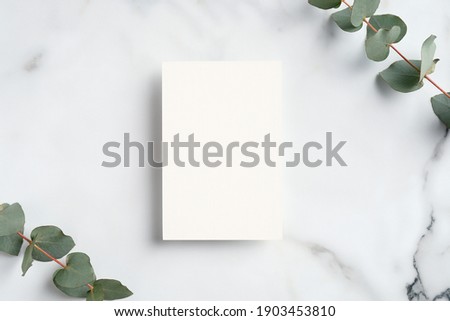Wedding invitation mockup. Blank paper card and eucalyptus branches on white marble table. Elegant rustic template.