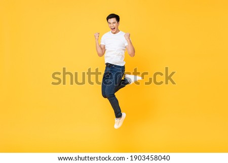 Happy energetic young Asian man jumping yelling and clenching fists isolated on yellow studio background, selective focus Royalty-Free Stock Photo #1903458040