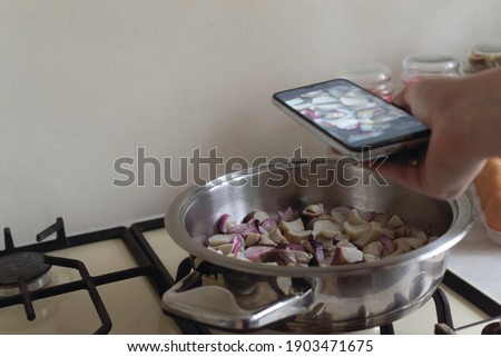 A woman photographs porcini mushrooms and red onions in a frying pan in her kitchen. Food blogger. Social media concept. Horizontal orientation. Copy space.