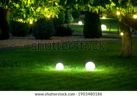 illumination garden light with electric ground lantern with ball diffuser lamp on meadow with garland of warm light bulbs on tree branch with leaves with landscaping, illuminate evening scene nobody. Royalty-Free Stock Photo #1903480186