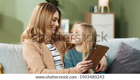 Happy mom with little daughter sitting hugging on sofa at home and watching cartoons online using tablet.