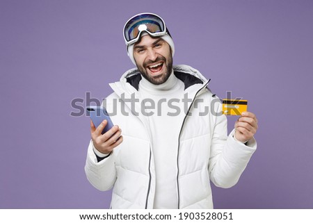 Funny skier man in white windbreaker jacket ski goggles mask using mobile phone hold credit bank card spend weekend winter in mountains isolated on purple background. People lifestyle hobby concept