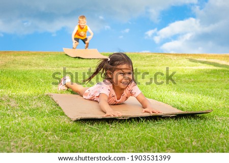 Children having fun on a summer day. Kids playing at the park sliding down grass hills on cardboard boxes.