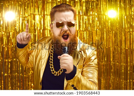 Funny singer in shiny golden jacket and gold chain holding microphone and singing songs at retro pop music concert. Happy bearded man performing at disco nightclub or enjoying time at karaoke party Royalty-Free Stock Photo #1903568443