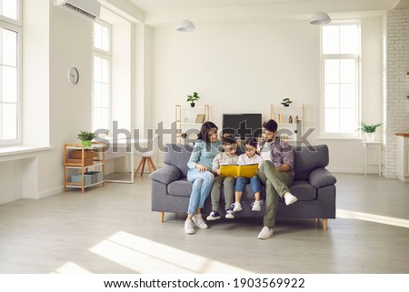 Happy people sitting together on sofa in modern interior of new house or studio apartment. Family with kids reading book on comfy gray couch in the living-room and enjoying quiet leisure time at home Royalty-Free Stock Photo #1903569922