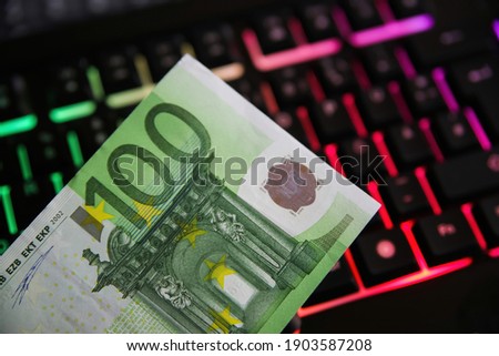 Closeup of isolated 100 euro paper money bank note, blurred illuminated computer keyboard background - online internet darknet trade concept (focus on banknote)