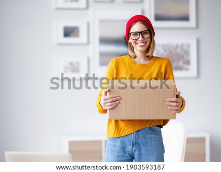 Delighted young female in trendy outfit smiling for camera and carrying cardboard box at home Royalty-Free Stock Photo #1903593187