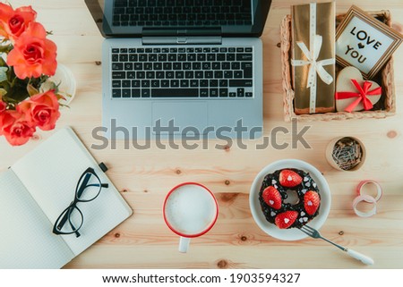 Top view working place with laptop, notebook, glasses and sweet Valentine's day surprise breakfast. Cup of latte coffee, bagel with strawberries and candy hearts, gift boxe, flowers for special one