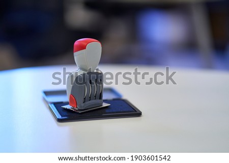 Office Stamp with Four Rolling Wheels, Resting on an Open Stamp Pad Filled with Blue Ink. Red Details on the Stamp Head and Body. White, Curved Office Desk. Blurred Background, Window Reflections Royalty-Free Stock Photo #1903601542