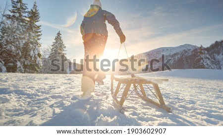 LOW ANGLE, LENS FLARE, CLOSE UP: Unrecognizable energetic woman runs up a snowy hill with her wooden sleigh. Excited female tourist rushes to the mountaintop to sleigh down the snow covered slope. Royalty-Free Stock Photo #1903602907