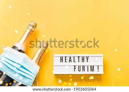 Healthy Purim written in lightbox, two champagne bottles, and medical mask on a yellow background. Flat lay of Purim Carnival celebration concept.