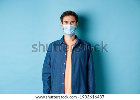 Covid-19 and healthcare concept. Smiling young man in medical mask looking healthy and happy, standing on blue background