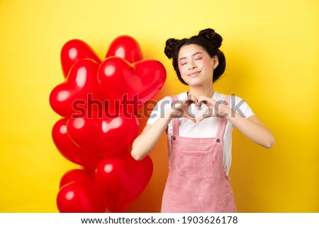 Valentines day concept. Cute asian girl dreaming of romance, close eyes and showing heart gesture, smiling happy, standing near red romantic balloons, yellow background