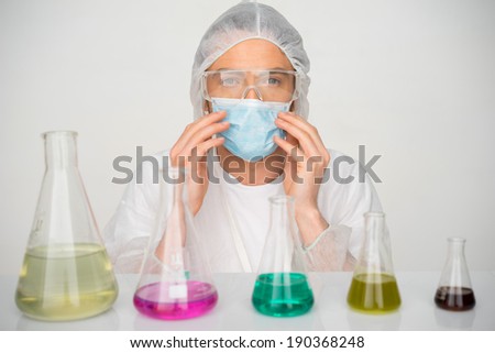 Male technician wearing a mask and cap working in a chemistry laboratory sitting behind a row of colorful chemical solutions in conical Erlenmeyer flasks
