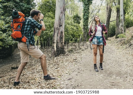 Caucasian female traveler posing for photo on road in forest and carrying backpacks. Young man holding camera and shooting her. Couple trekking together on nature. Tourism and vacation concept