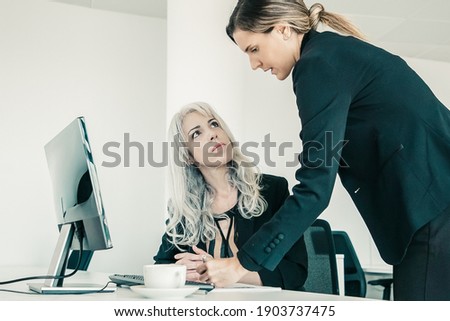 Project manager affixing signature on employees report. Female business colleagues sitting and standing at workplace with monitor and coffee cup. Business communication concept