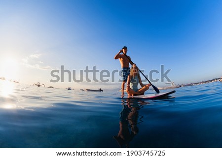 Young happy couple have fun on stand up paddleboard. Active paddle boarder paddling by sunset sea. Healthy lifestyle. Water sport, SUP surfing tour in adventure camp on family summer beach vacation