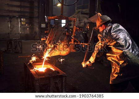Foundry worker in protective suit and hardhat filling mold with hot molten iron to make parts for industry. Royalty-Free Stock Photo #1903764385