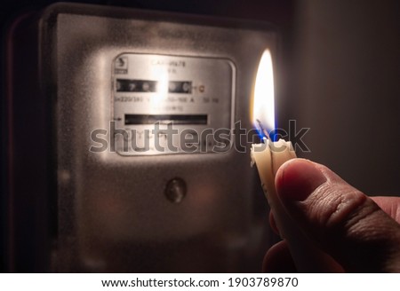 A man's hand with candle in complete darkness near an electricity meter at home. Power outage, blackout, non-payment of electricity bill concept. Royalty-Free Stock Photo #1903789870