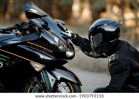 Motorcycle driver in leather suit and helmet sits in front of the motorcycle alone and put his hands on the headlights Royalty-Free Stock Photo #1903793110