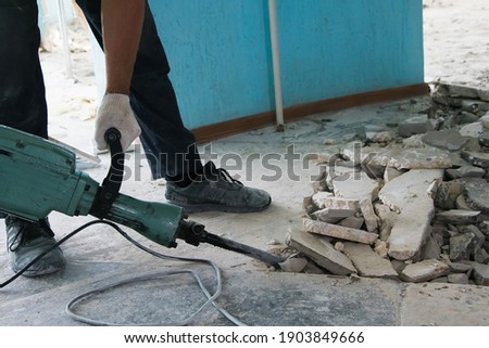 A worker uses a jackhammer to remove a layer of old cement screed inside the repaired room. Construction Royalty-Free Stock Photo #1903849666