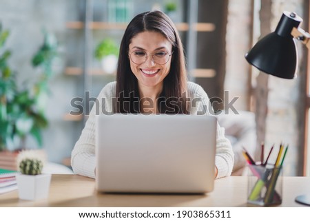 Photo of worker lady sit desk work computer look screen beaming smile wear eyeglasses white pullover in living room home indoors