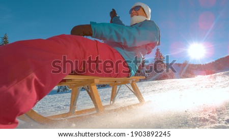 LOW ANGLE, LENS FLARE, CLOSE UP: Cheerful Caucasian woman on carefree winter vacation in scenic Kranjska Gora sleds down snowy hill on a picturesque snowy day. Happy female tourist riding a sleigh.