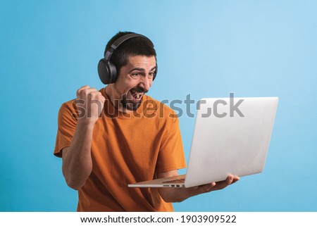 Young handsome man with gamer headphone set isolated over blue background using laptop computer playing a game and celebrating victory.