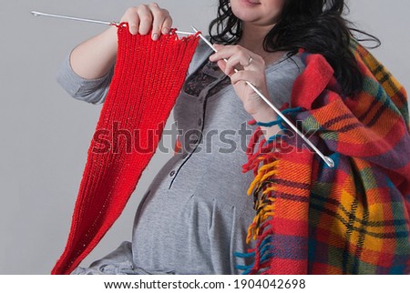 A young pregnant woman in a gray home dress, covered with a woolen checkered plaid, holds knitting needles in her hands and knits a red scarf. No face visible. Close. Studio.