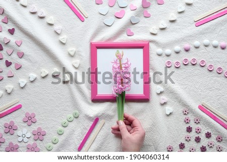 Abstract Springtime background. Hand holding pink pearl hyacinth flower in pink picture frame. Flat lay with hearts, candy, buttons, small decor arranged on textile background. Off white tablecloth.