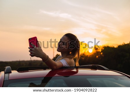 Beautiful young woman taking a selfie while enjoying a ride - Woman takes selfie on the roof of a car at sunset .