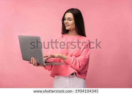 Photo of attractive lady in glasses having online meeting, holds laptop and works online. wear casual pink t-shirt white pants isolated pink color background.