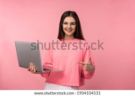 Photo of attractive lady in glasses, smiles and has online meeting, holds laptop and works online. wears casual pink t-shirt white pants isolated pink color background. Online study.