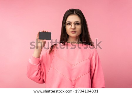 Photo of attractive lady in glasses holds credit card, wears casual pink t-shirt white pants, isolated pink color background.