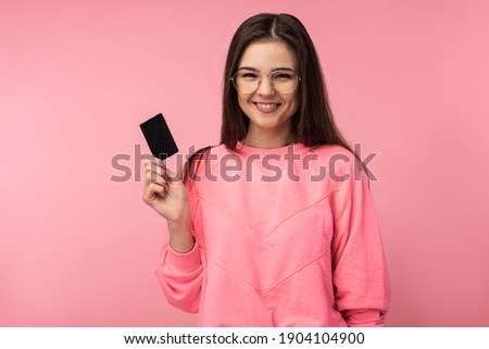 Photo of attractive lady in glasses, smiles and holds credit card, wears casual pink t-shirt white pants isolated pink color background.