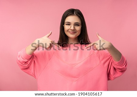 Photo of attractive woman in glasses smiles and points at something, wear casual pink t-shirt white pants isolated pink color background