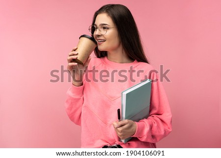 Photo of attractive lady in glasses holds book and drinks coffee. Student works or studies online. Wear casual pink t-shirt white pants isolated pink color background