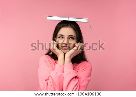 Photo of attractive woman in glasses covers her head by book. Bewildered student works studies online. Wears casual pink t-shirt white pants isolated pink color background